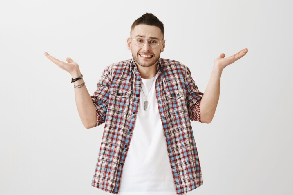 Confused embarrassed attractive adult man with bristle, wearing transparent glasses, shrugging with raised palms, smiling nervously, feeling guilty over gray background. Oops, my bad. It was my fault