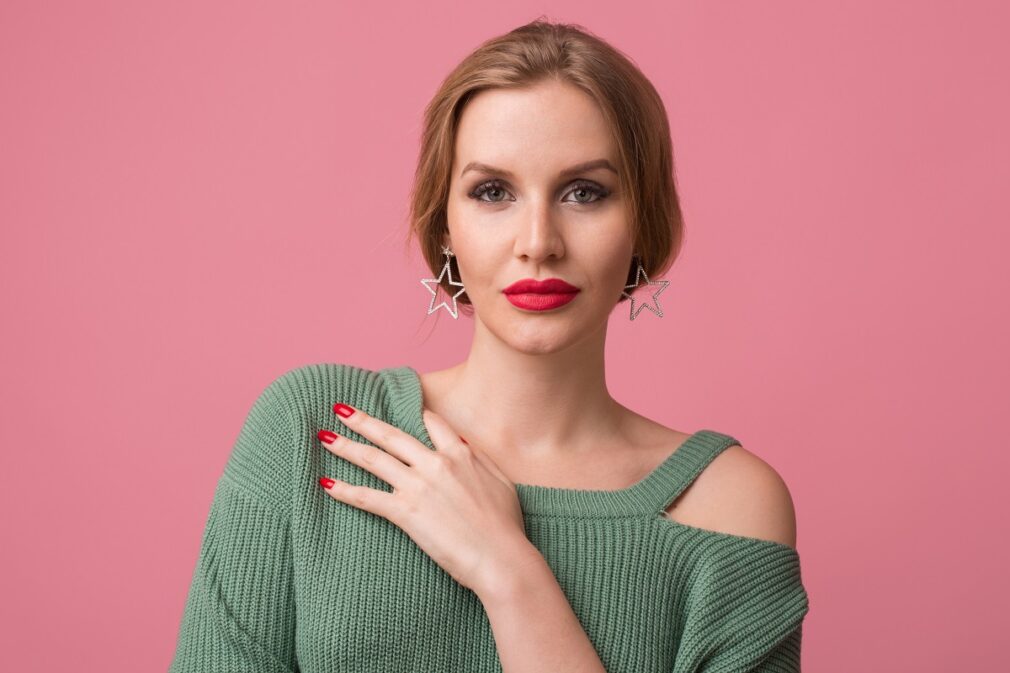 close-up portrait of young sexy attractive woman, stylish make-up, red lips, green sweater, model posing in studio, isolated, pink background, earrings, looking in camera