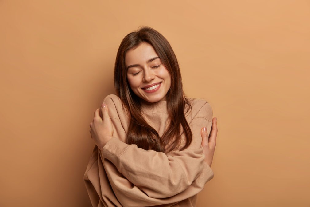 I love myself. Pretty smiling woman embraces herself gently, expresses self love, keeps eyes closed with pleasure, feels comfortable and fullfilled, being egoistic person, isolated on brown background